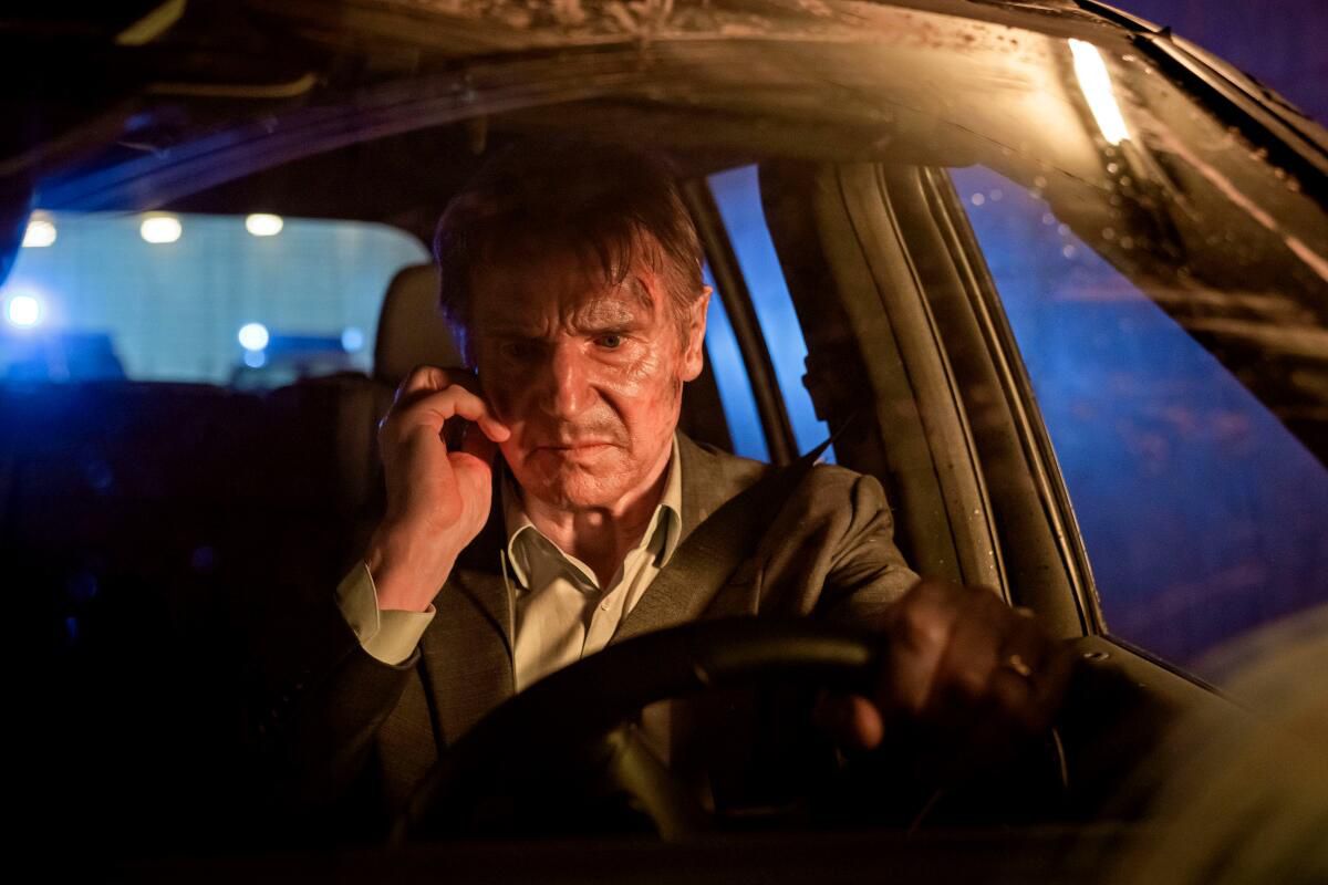 Liam Neeson, synbart rufsig, sitter bakom ratten i en bil med en mobiltelefon bredvid örat i Retribution.