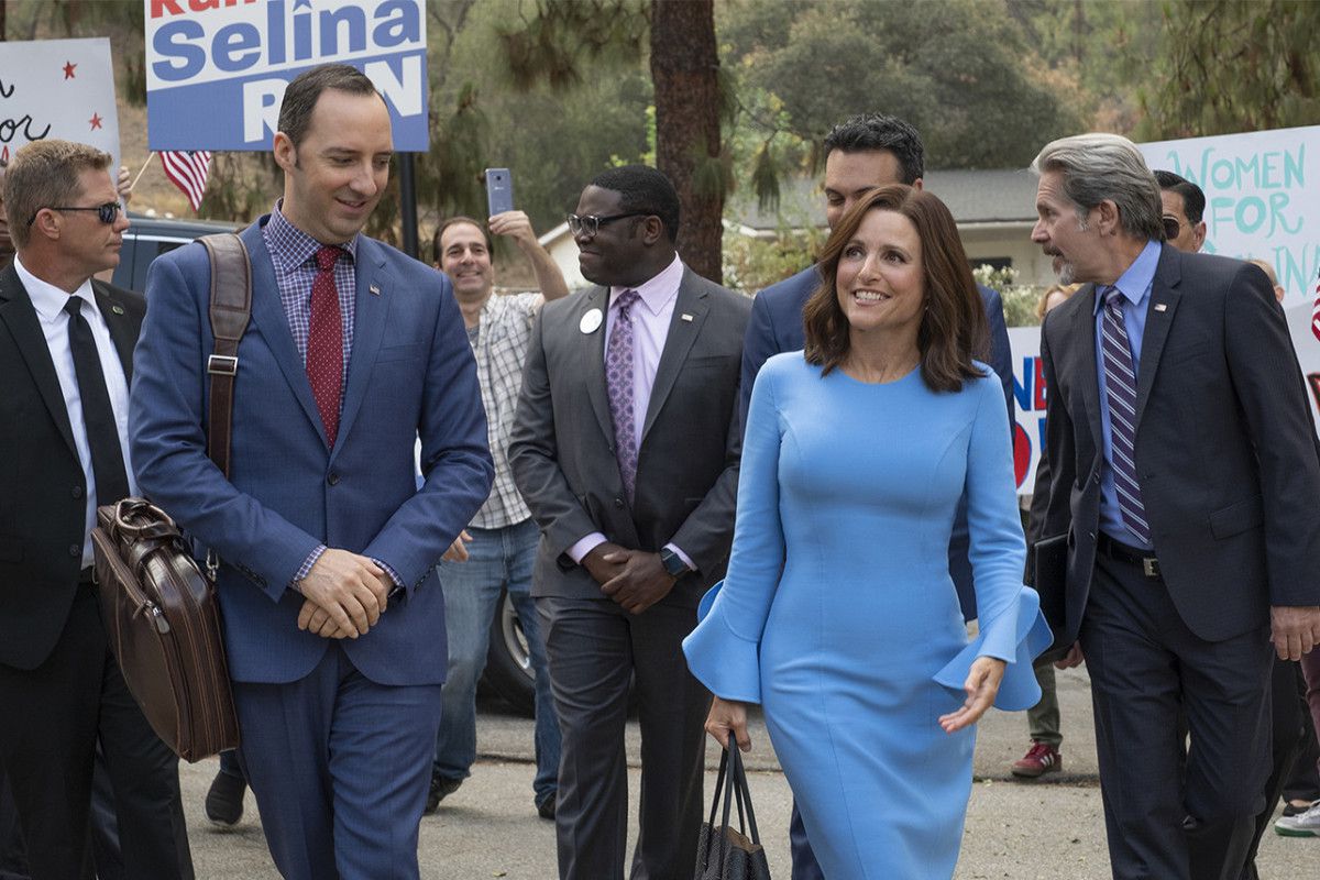 Julia Louis-Dreyfus, wearing a blue dress, walks with campaign posters around her, along with Tony Hale, Sam Richardson, and other supporting staff.