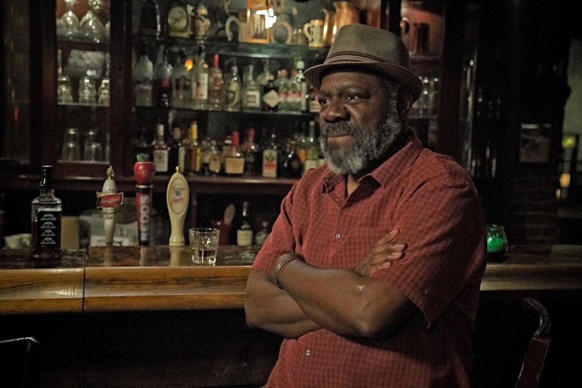Frankie Faison, wearing a hat and a red polo, crosses his arms while sitting at a bar in Banshee.