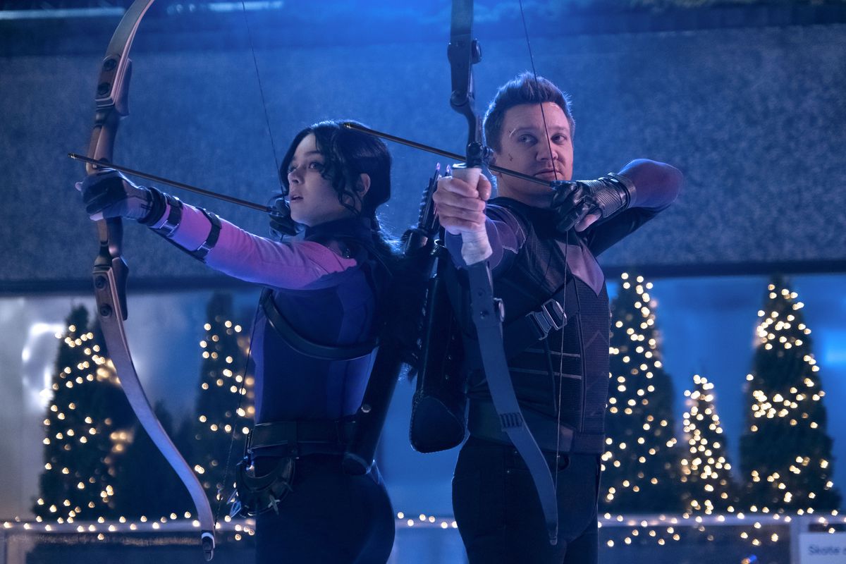 Kate Bishop and Clint from Avengers in Marvel’s Hawkeye standing back to back with bows and arrows in 30 Rock Plaza