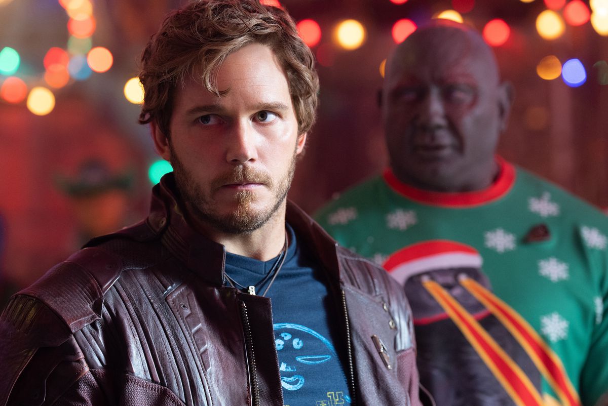 Human hero Peter Quill/Star-Lord looks frustrated in the foreground as his teammate Drax the Destroyer (Dave Bautista, in purple makeup, bright-red scarification designs, and a comically garish Christmas sweater depicting a creature with laser-eyes) smiles in the background in The Guardians of the Galaxy Holiday Special