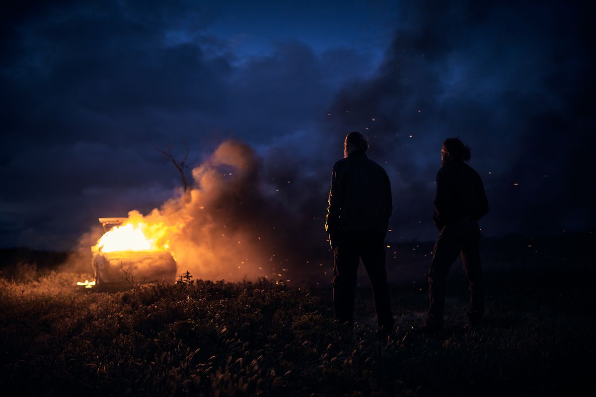 Two man stand in a field at night watching acar erupt in a blaze of fire.
