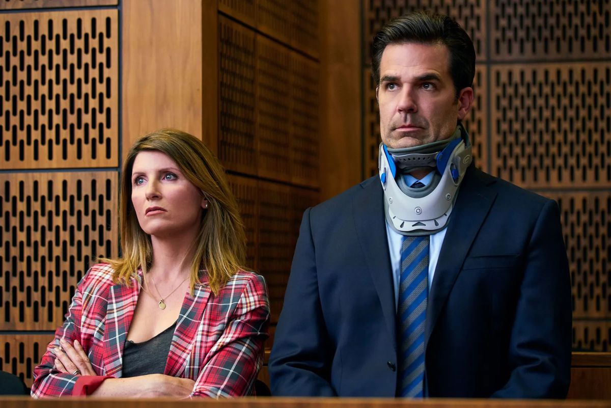 Rob Delaney, wearing a suit and neck brace, stands next to Sharon Horgan in what looks like a courtroom in Catastrophe.