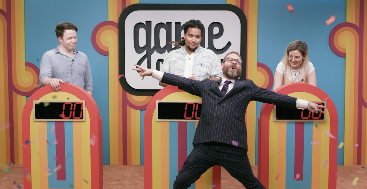 Sam Reich, host of Game Changer, celebrates effusively in front of the show’s iconic podiums. Three contestants — Brennan Lee Mulligan, Lou Wilson, and Siobhan Thompson — stand behind the podiums, radiating frustrated tolerance.