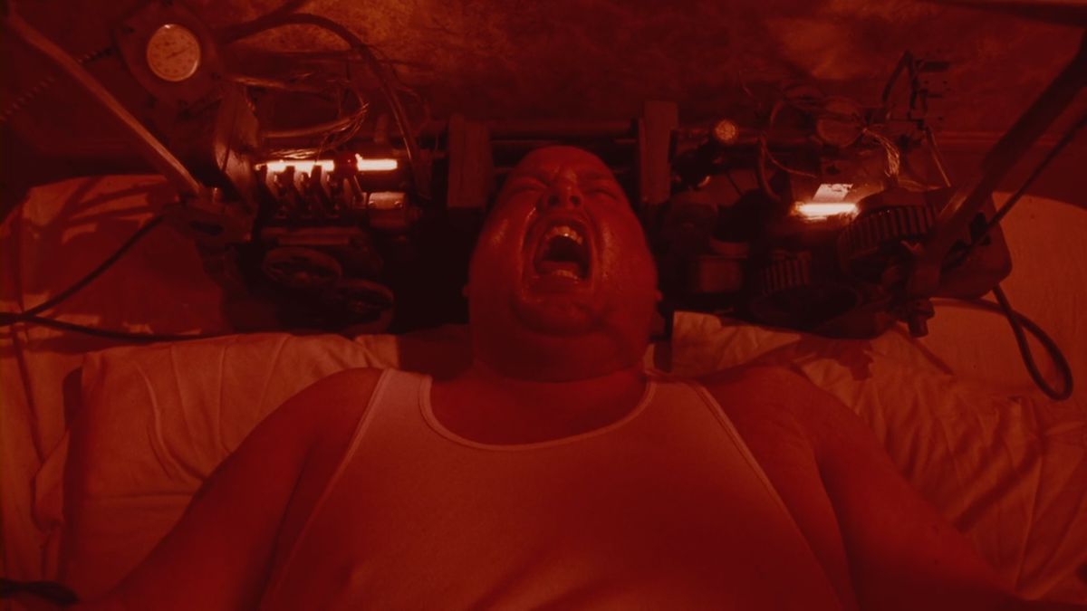 En man som ligger i sängen iförd linne skriker med en udda grej runt huvudet, i en rödtonad bild från Saw IV.