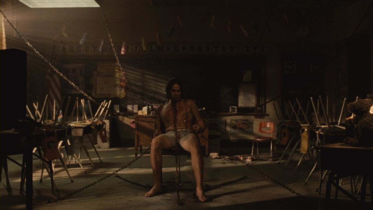 En man, endast iklädd underkläder, sitter i ett tomt klassrum.  Han har kedjor fastkrokade på sitt kött.  Detta är i Saw III, förstås.