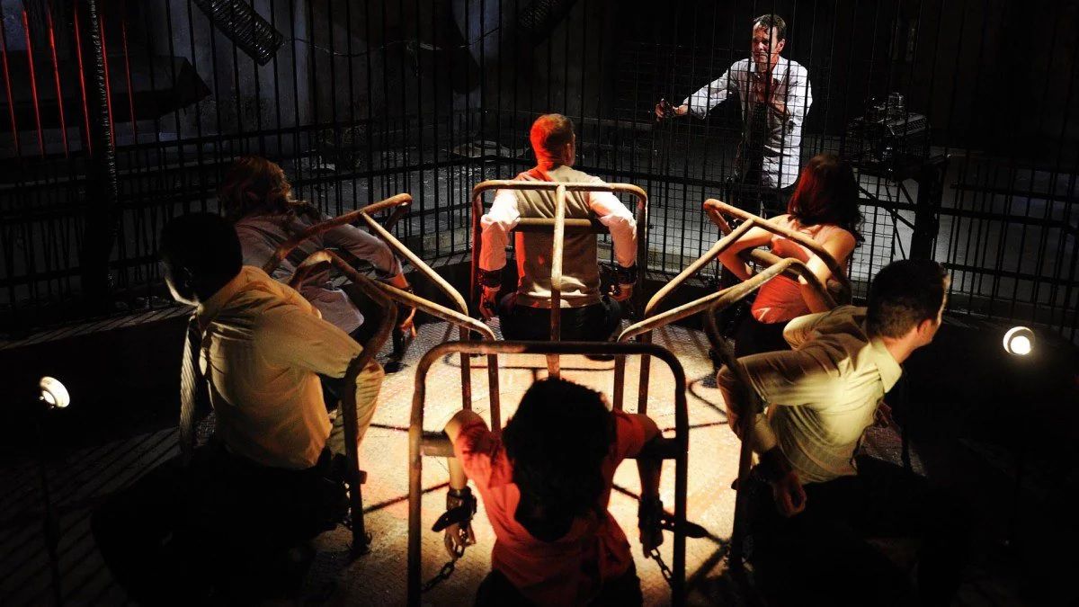 En grupp människor handfängslade i en karusell tittar på en person utanför buren de är instängda i i SAW VI.