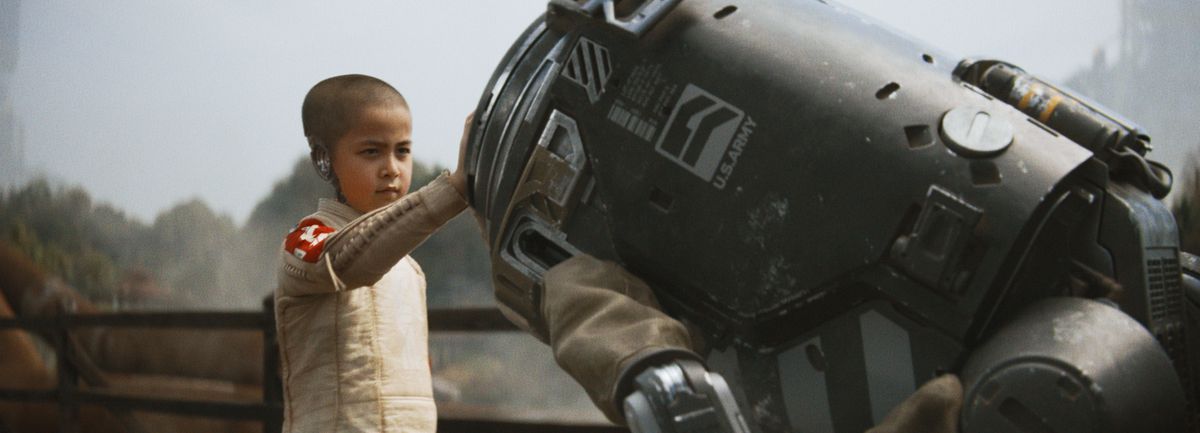 en ung cyborgtjej med ett rakat huvud lägger sin hand på en papperskorgsliknande robot som böjer sig för henne i The Creator