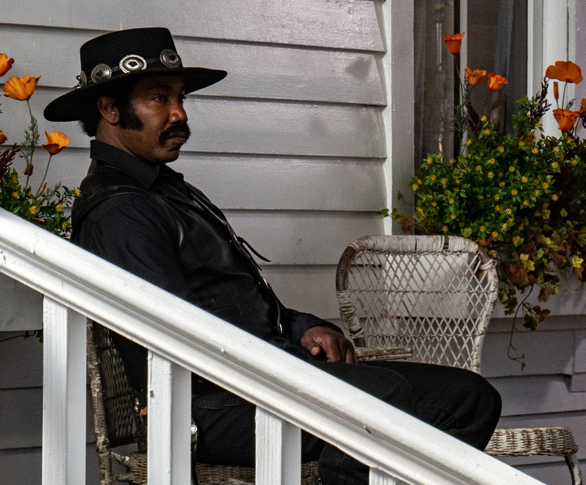 Michael Jai White som Outlaw Johnny Black sätter sig på en solstol, klädd i helsvart med en svart cowboyhatt, långa sidobrännskador och en tjock mustasch.
