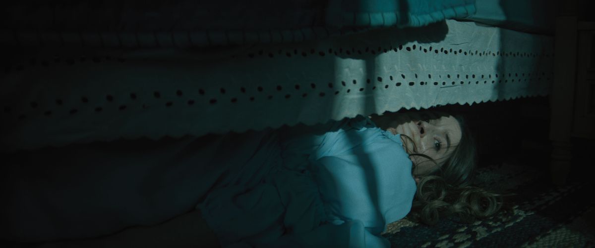Brynn Adams (Kaitlyn Dever), en långhårig ung kvinna i blått nattlinne, gömmer sig under sin säng i mörkret med handen klappad över munnen i skräckfilmen No One Will Save You