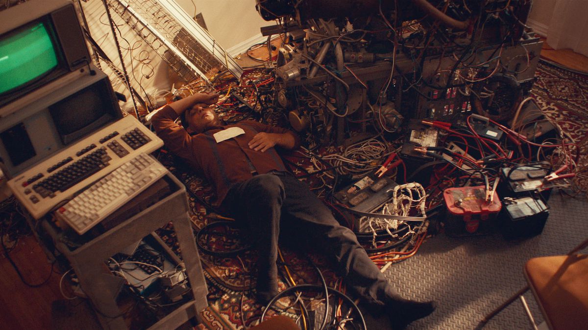 Medelålders uppfinnare Jabir (Payman Maadi) spretar på rygg på golvet med ena armen slängd över huvudet, omgiven av sladdar, startkablar, bilbatterier, en uråldrig grön skärm och tangentbord och annan gammaldags teknik i Aporia