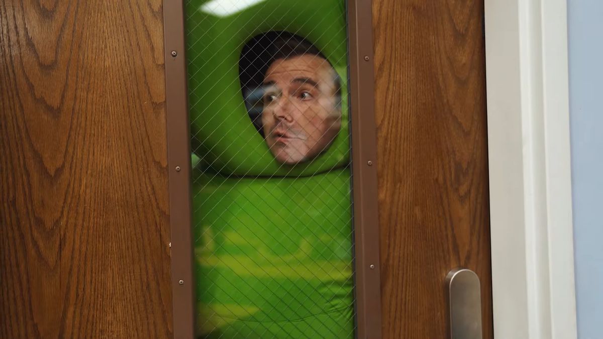 En man klädd som en schackpjäs trycker sitt ansikte mot fönsterrutan i en klassrumsdörr.