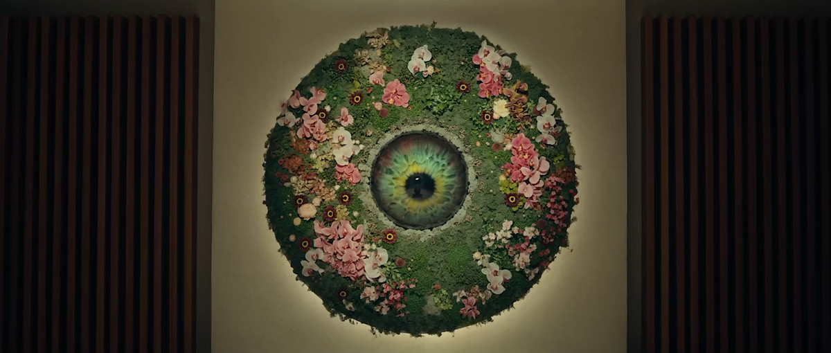 AI-terapeuten från The Pod Generation, en enorm grön ögonglob inbyggd i en vägg, omgiven av en cirkulär skärm av grönt gräs och rosa och vita blommor