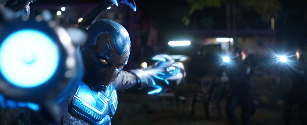 Blue Beetle poserar med armarna i form av laserkanoner pekande både direkt mot och bort från kameran i filmen Blue Beetle.