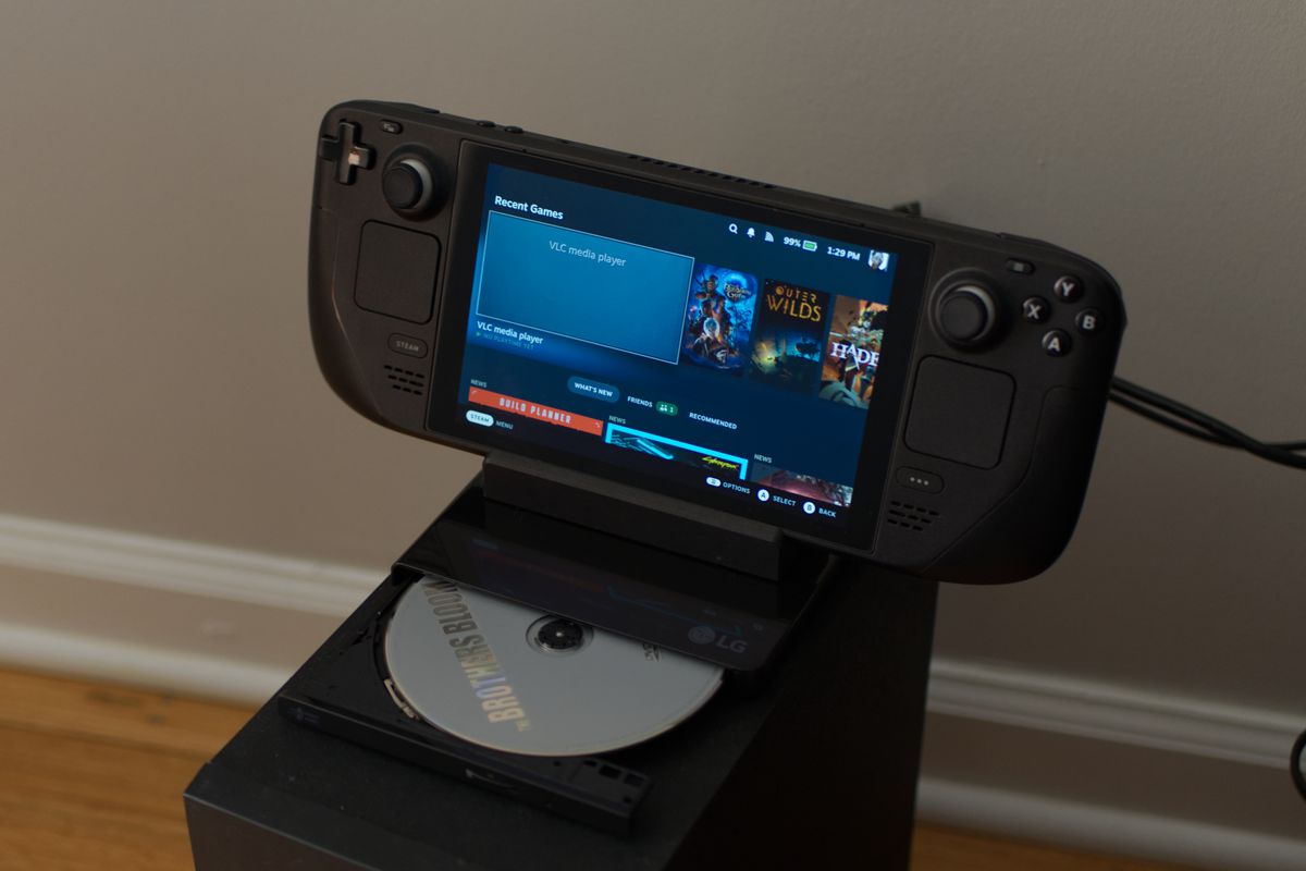 En bild som visar den bärbara Steam Deck-konsolen inkapslad i sin TV-docka.  Däcket och dess docka sitter ovanpå en extern DVD-spelare som är ansluten till dockan.