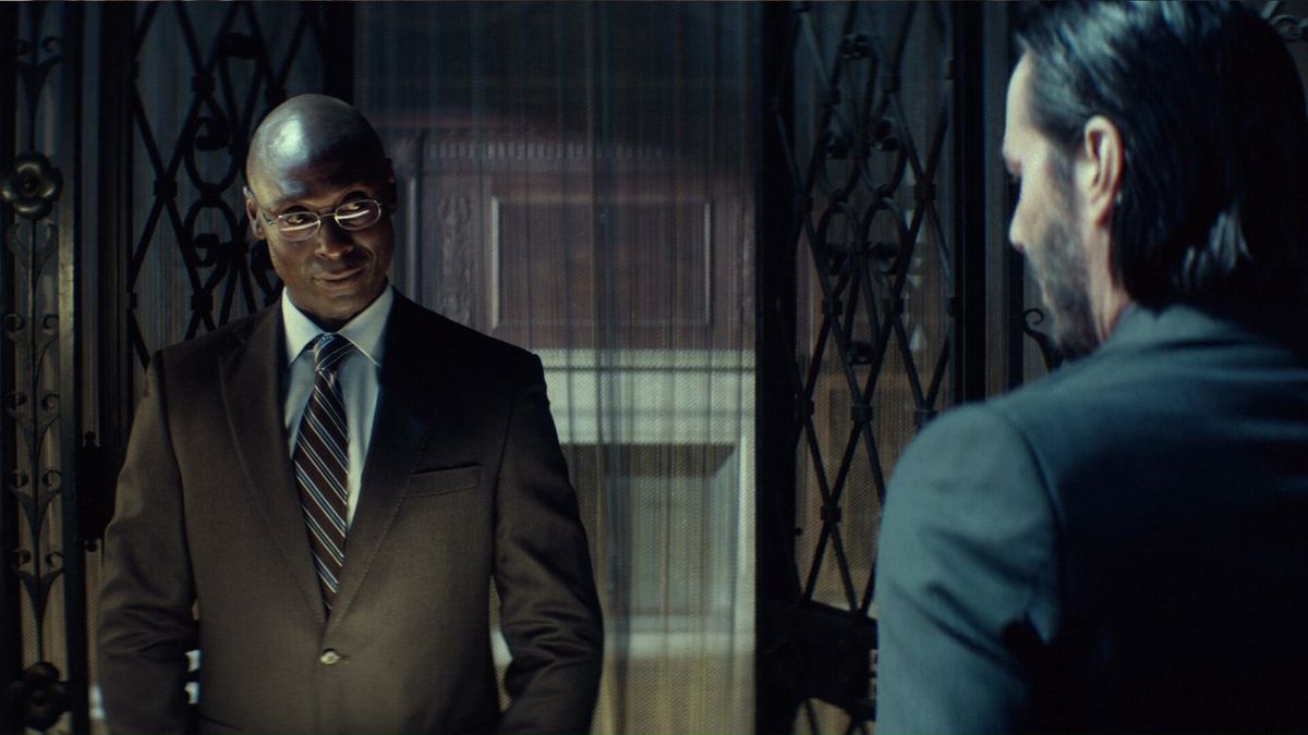 Lance Reddick som concierge Charon, i en snygg brun kostym, hälsar Keanu Reeves som John Wick vid incheckningsdisken på Continental hotel i John Wick: Kapitel 3 – Parabellum
