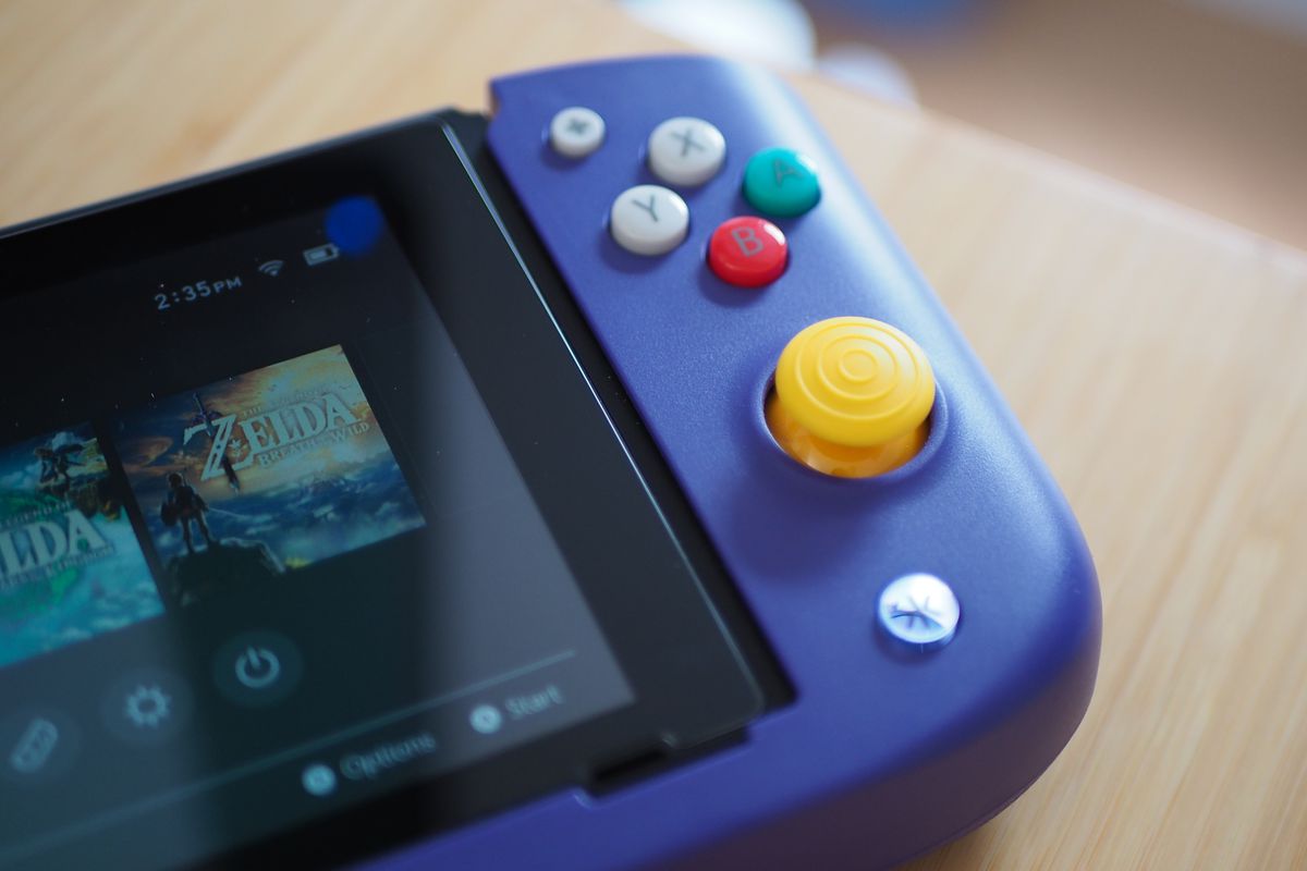 Ett foto på höger sida av Crkd Nitro Deck, en Nintendo Switch-hållare som har alla konsolens kontroller och portar.  Den är lila med färgade joysticks och knappar, som efterliknar Nintendo Gamecube.