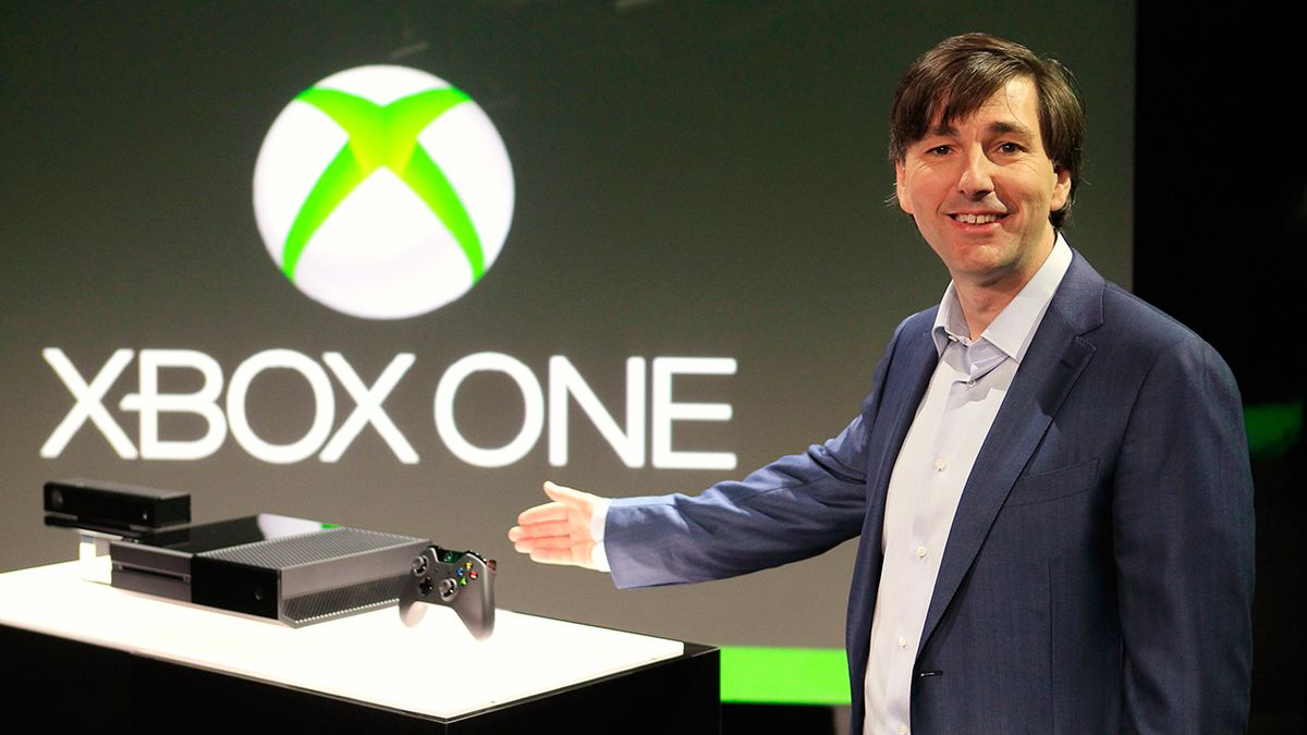 En disketthårig chef, Don Mattrick, ler medan han gör en gest mot en Xbox One-konsol framför en stor Xbox One-logotyp