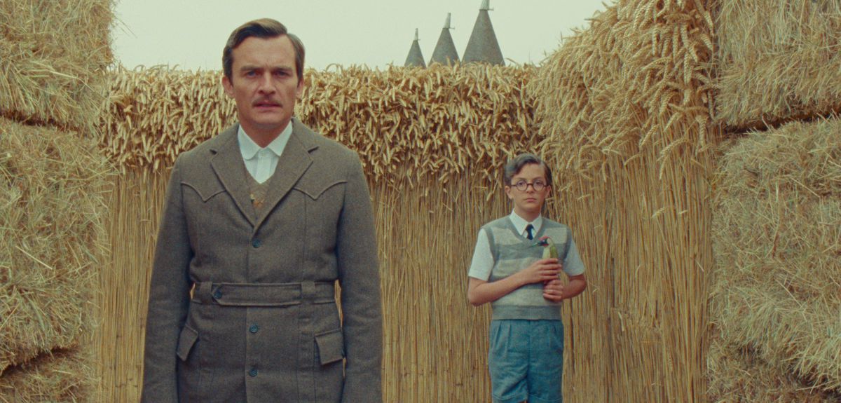 Rupert Friend as the narrator stands in front of schoolboy Peter Watson (Asa Jennings), a small kid in blue wearing round glasses and clutching a book. They’re framed by walls of hay bales and vertical dried hay, in a scene from Netflix’s The Swan