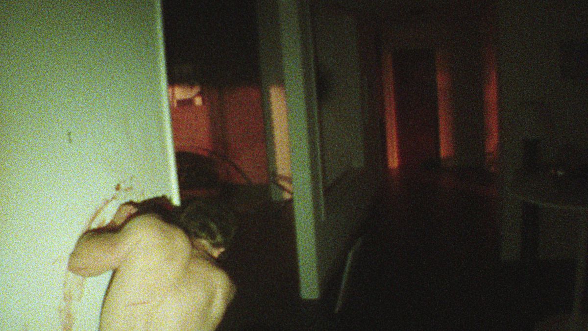 En man utan bar överkropp, sedd bakifrån, hukar i en hall i en mörk, kornigt skjuten lägenhet, lutad mot en smutsig grönaktig vägg och smetar blod på den i V/H/S/85-segmentet Dreamkill