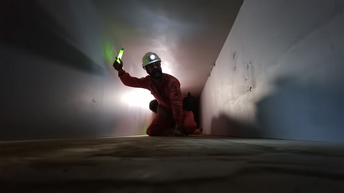 En man i orange byggnadsarbetarskrud och en hjälm med en lampa på håller upp en grön glowstick när han kryper genom en vit betongtunnel i en mycket kornig scen från V/H/S/85-segmentet 
