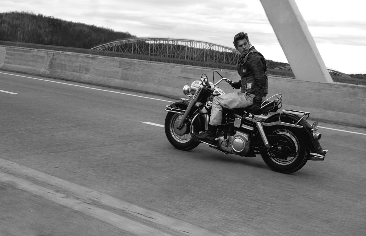 Austin Butler, i svart och vitt, åker sin Harley-Davidson över en bro med en hand och tittar tillbaka över axeln på ett väldigt coolt sätt i The Bikeriders