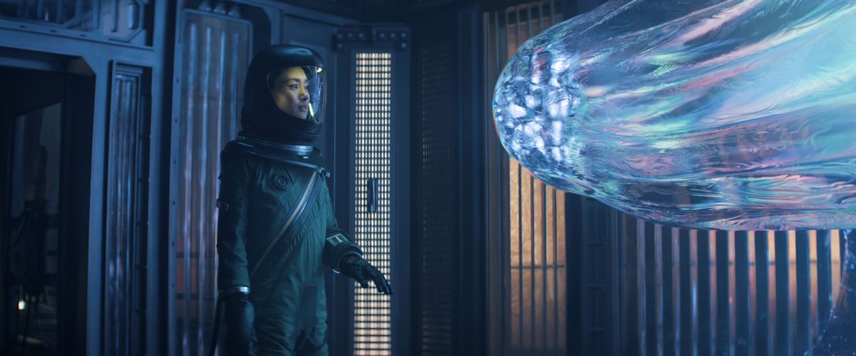 Mitsuki Yamato (Shioli Kutsuna) står i en rymddräkt i ett rum med en glob-liknande Entitet som lyser och rör sig närmare henne