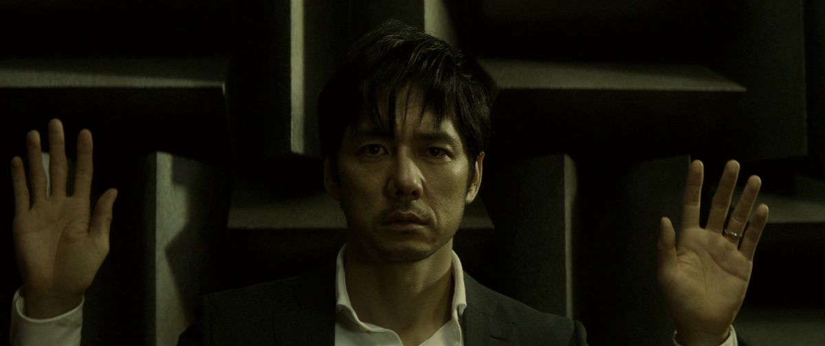 En karaktär från Kiyoshi Kurosawas film Creepy står med händerna upp mot en konstigt mönstrad bakgrund