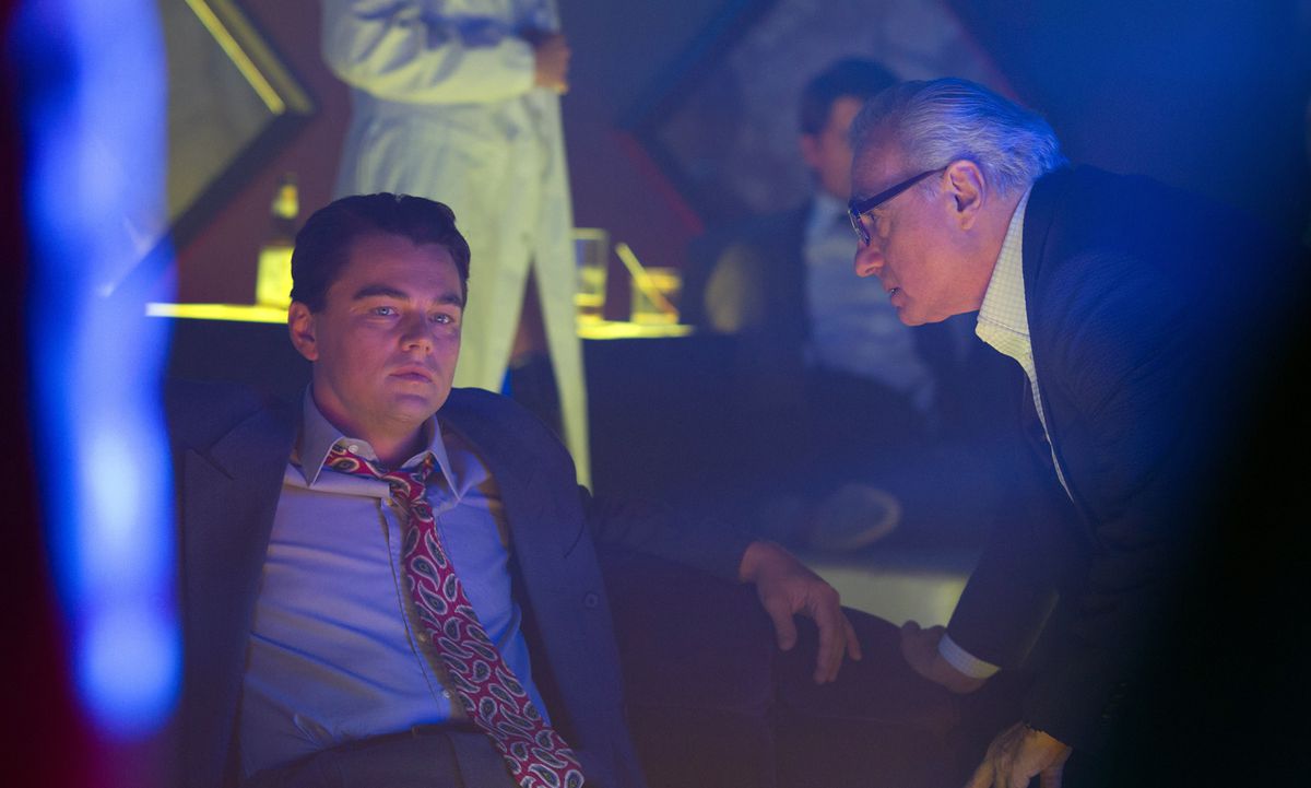 Martin Scorsese bends over to speak to Leonardo DiCaprio, who’s sitting on a couch in a dim, blue-light space, wearing in a suit and tie as Jordan Belfort on the set of The Wolf of Wall Street