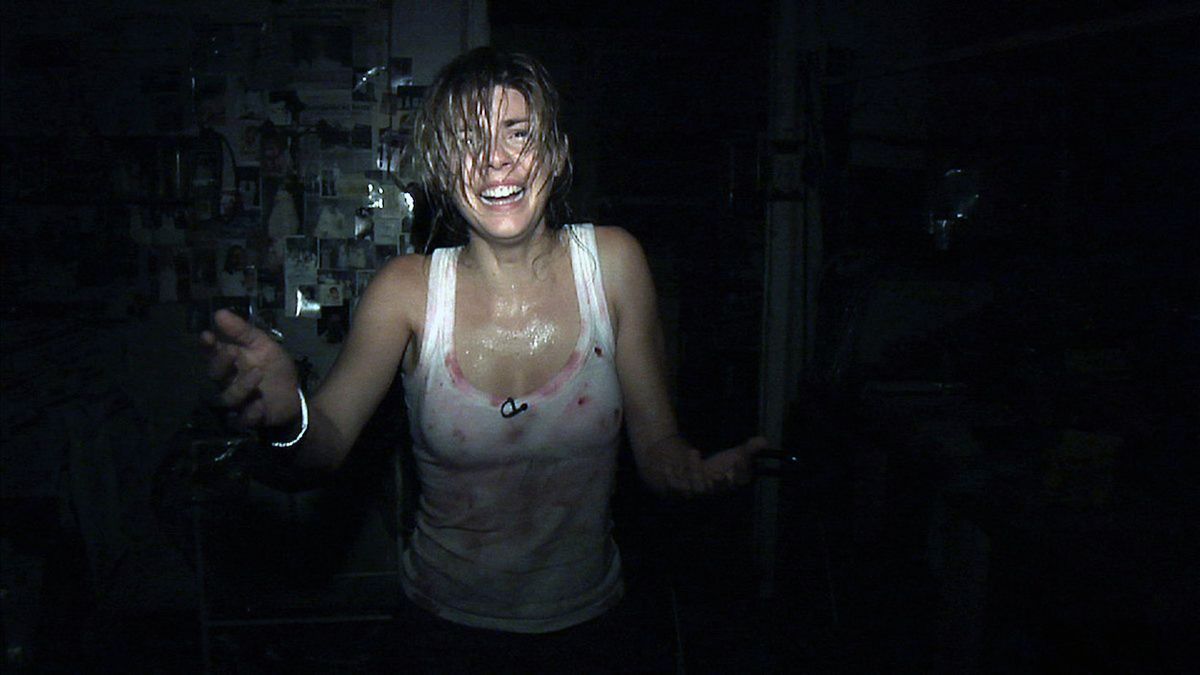 En gråtande kvinna i en vit linne prickad med blod står i ett mörkt rum upplyst av en kameralampa i REC.