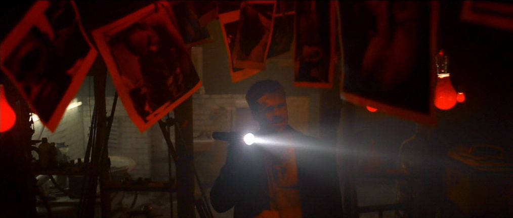 Brad Pitt som detektiv Mills lyser med en ficklampa i ett mörkt rum upplyst av röda glödlampor och dekorerat med foton i Seven.