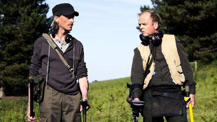 Mackenzie Crook and Toby Jones stand in a field with their metal detectors in Detectorists