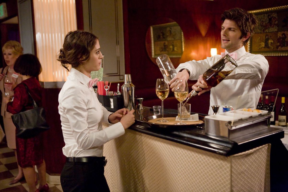 Adam Scott pours a drink while talking to Lizzy Caplan across the bar in Party Down. Both wear their catering uniforms