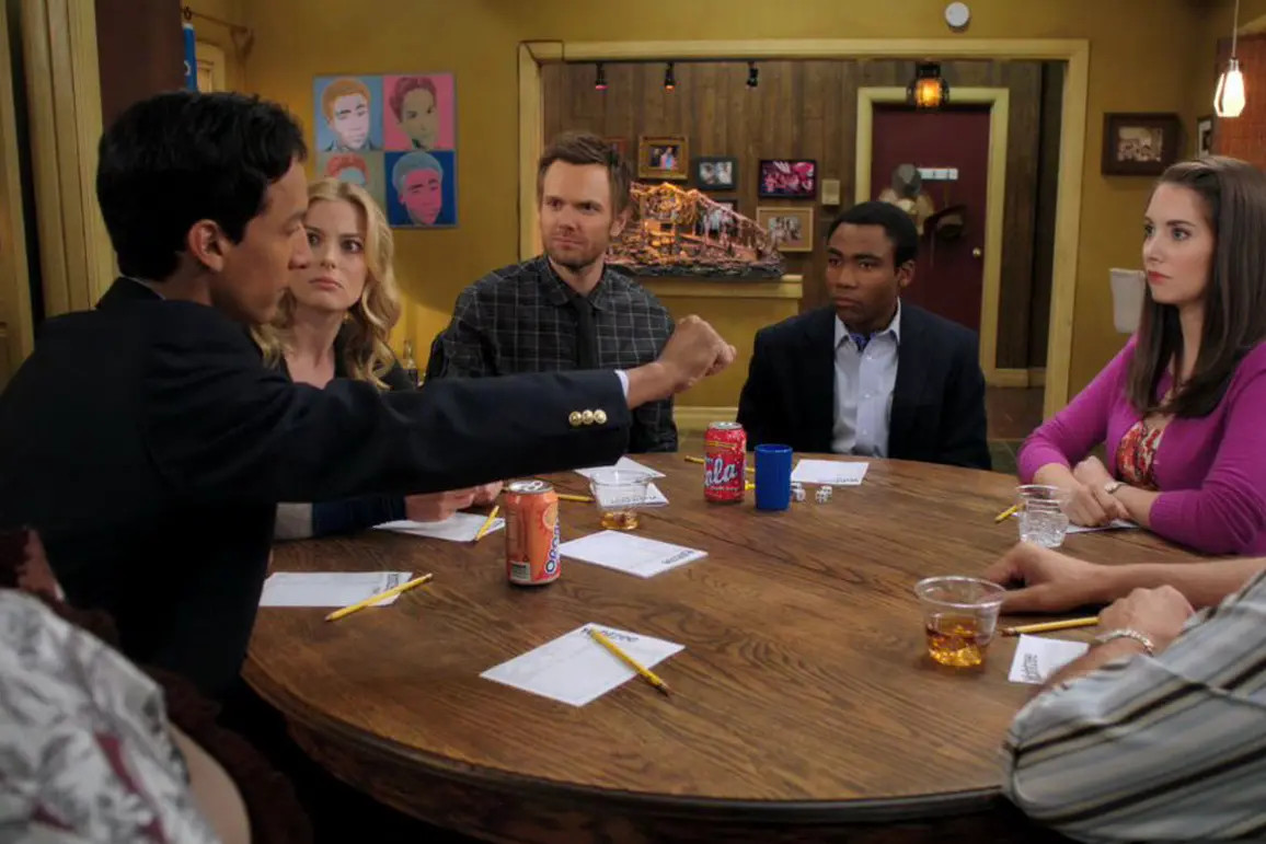 Danny Pudi catches a die in midair, as the rest of the main cast of Community stares at him around a table