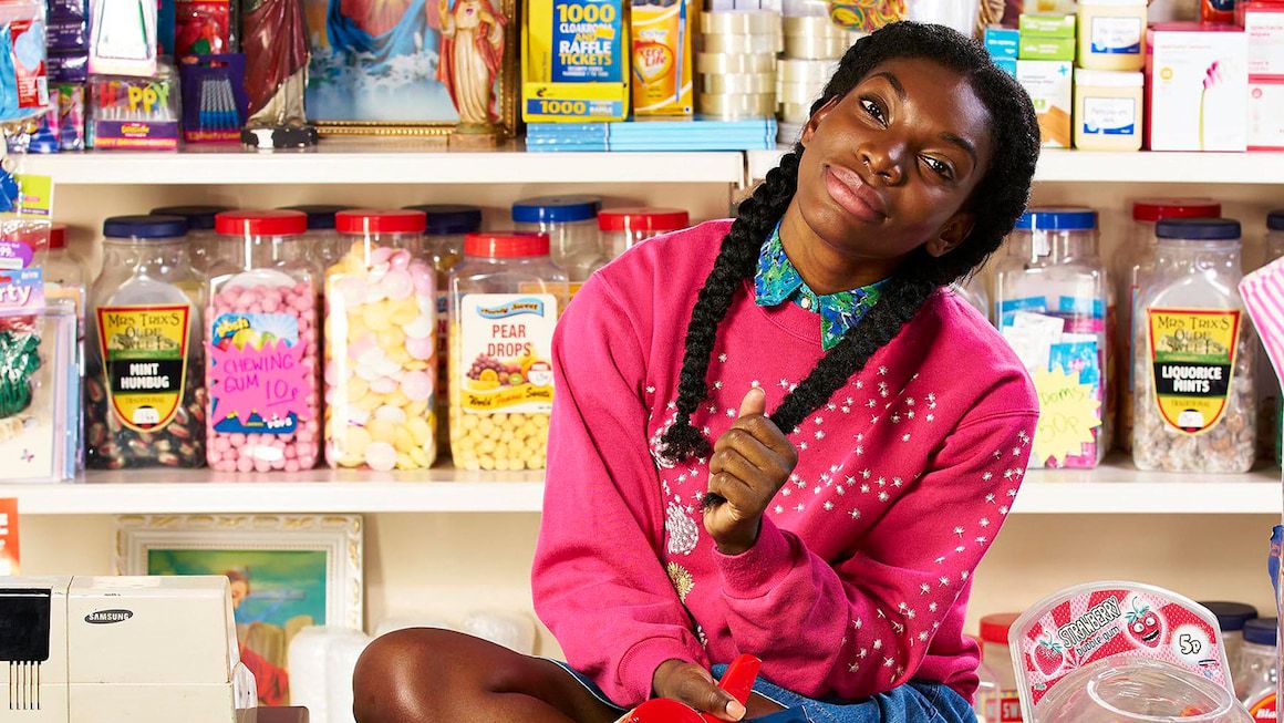 Michaela Coel wears a pink sweater and holds onto her braided hair, with tons of candy behind her, in Chewing Gum.