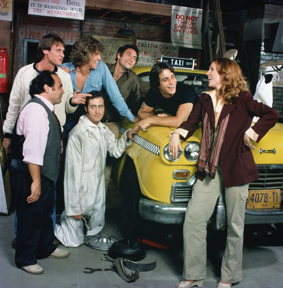 The cast of Taxi laugh in front of a taxi cab, with tools on the ground in front of it.