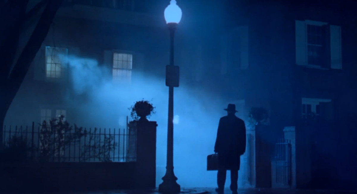 En man i hatt och kappa, bärande en portfölj, står siluetterad i det blå ljuset från en gatlykta i dimman som dyker upp från det upplysta andra våningsfönstret i ett hus i en scen från Exorcisten