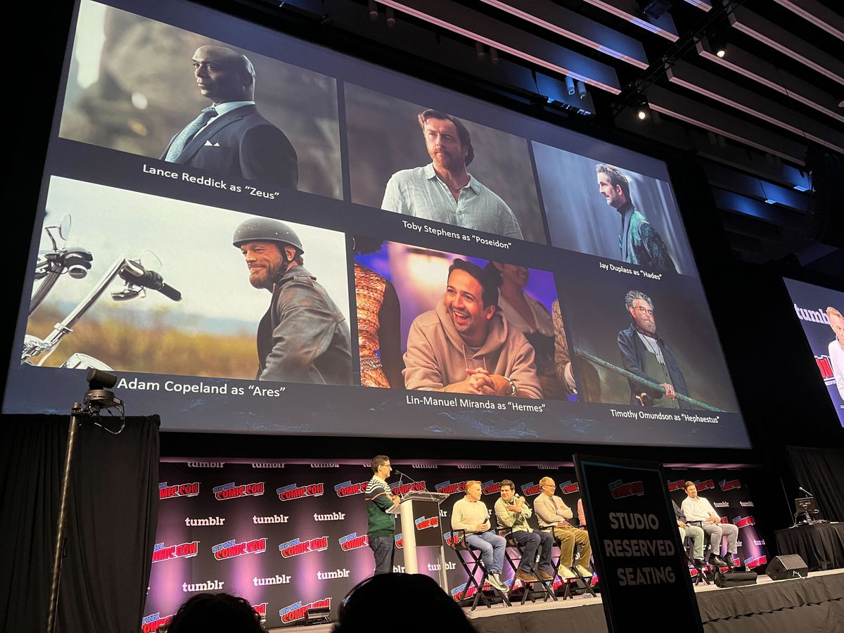 Ett foto av scenen på NYCC med Percy Jackson-teamet sittande i stolar framför en stor skärm som har bilder på de vuxna skådespelarna och deras titlar: 