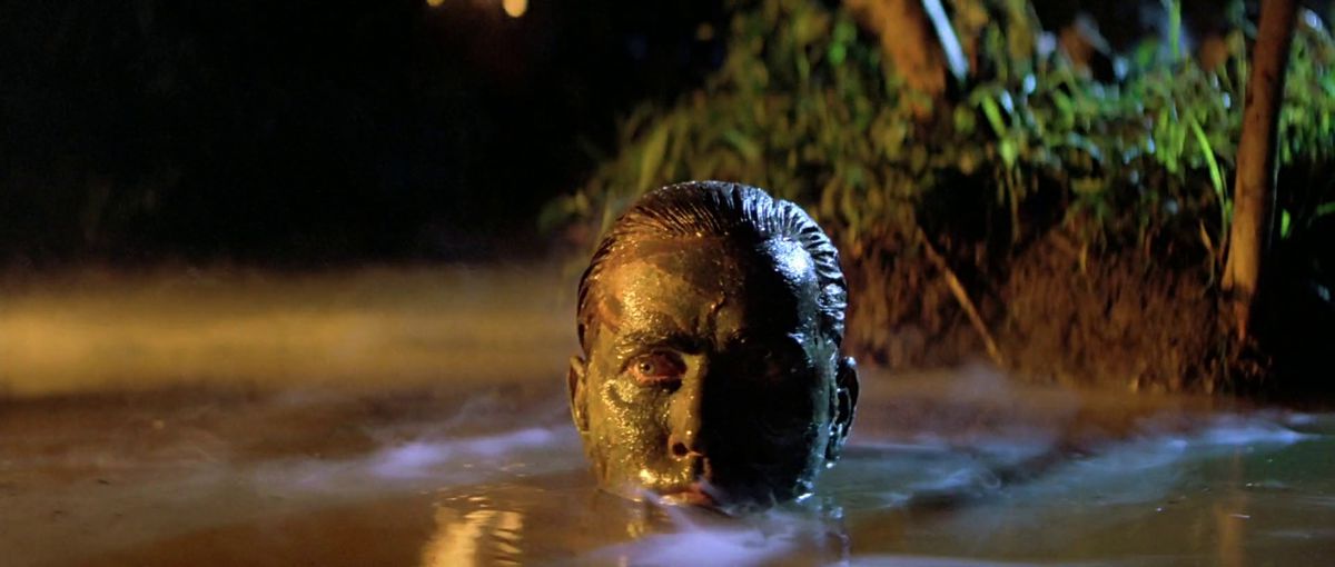 Martin Sheen som Benjamin Willard bär ansiktskamouflage och dyker upp ur en vattenmassa draperad i månbelysta dimma i Apocalypse Now.