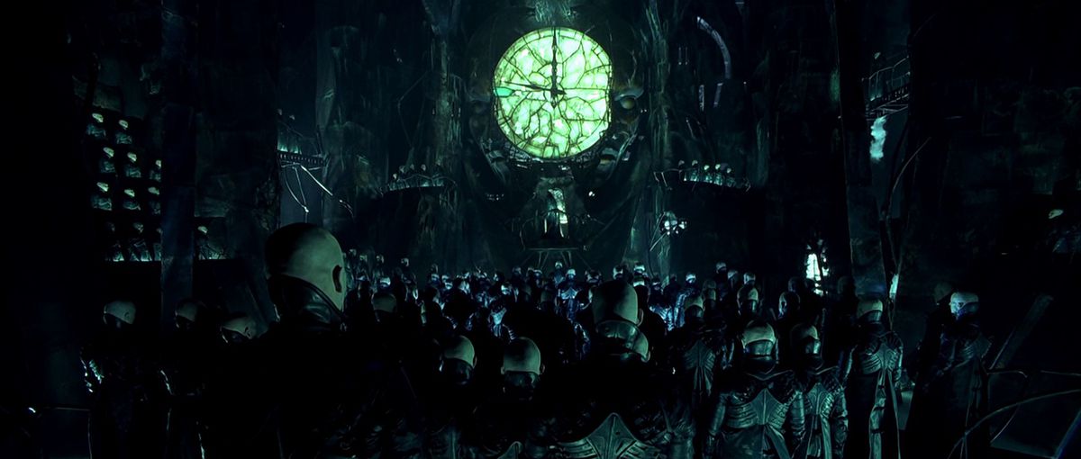 En skara skalliga män i läderkläder står mitt i ett stort atrium med en sjukt grön urtavla som sticker ut ur en staty i Dark City.
