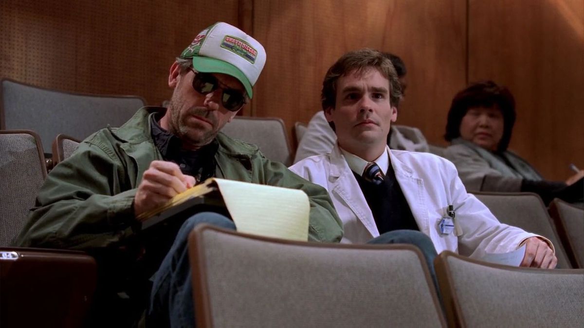 Hugh Laurie som Dr. House, klädd i solglasögon och en Gravedigger-keps, sitter bredvid Wilson i en föreläsning