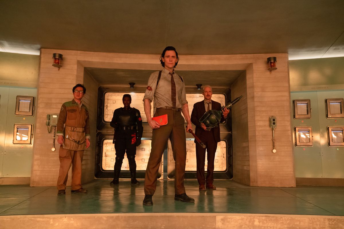 Loki står med en orange handbok i handen med medlemmarna i TVA, inklusive agent Mobius och Ouroboros, bakom honom.