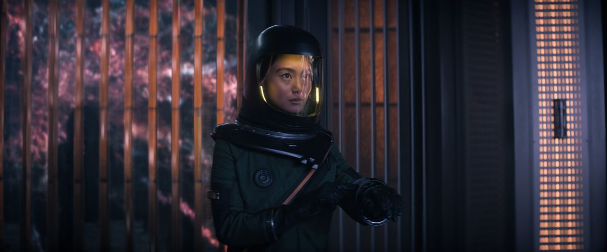 Mitsuki Yamato (Shioli Kutsuna) i en rymddräkt som håller upp händerna, med Entiteten reflekterad i hennes visir och ett fönster bakom sig