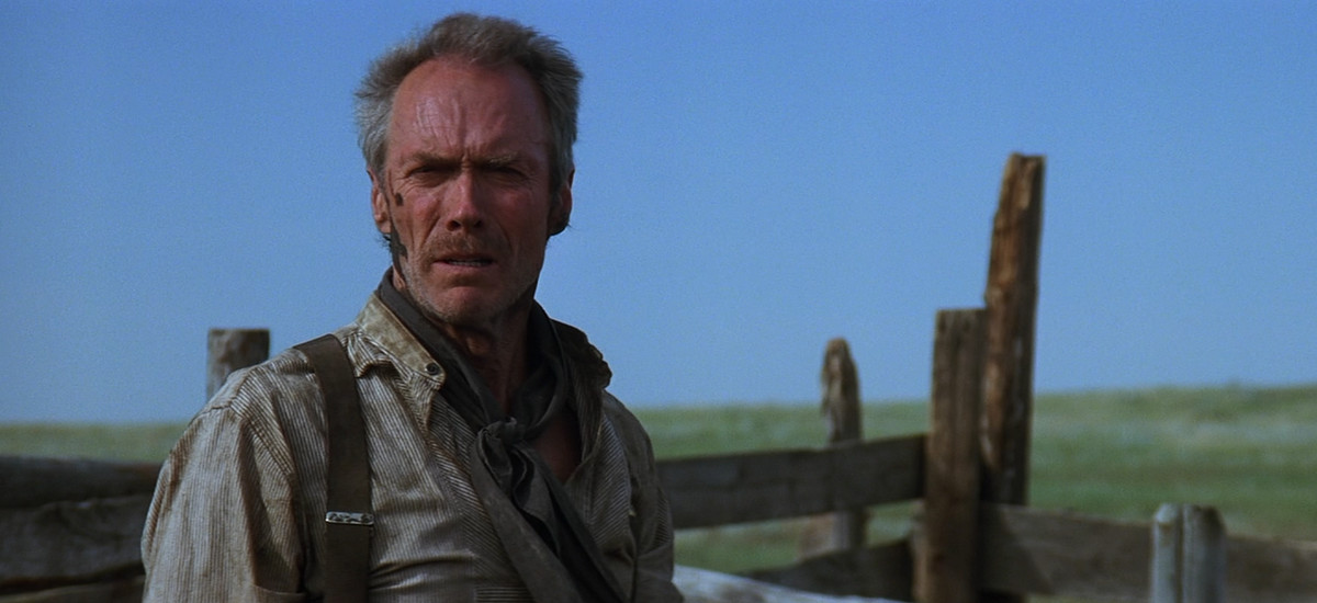 Clint Eastwood har smuts i ansiktet och ser bedrövad ut i Unforgiven, med horisonten och ett stort grönt fält bakom sig.