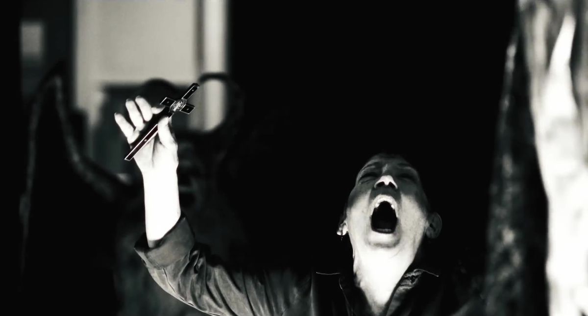 Ann Dowd i The Exorcist: Troende som håller upp ett kors och skriker i ett mörkt rum, med den mycket vaga skuggan av en bevingad figur i silhuett bakom henne