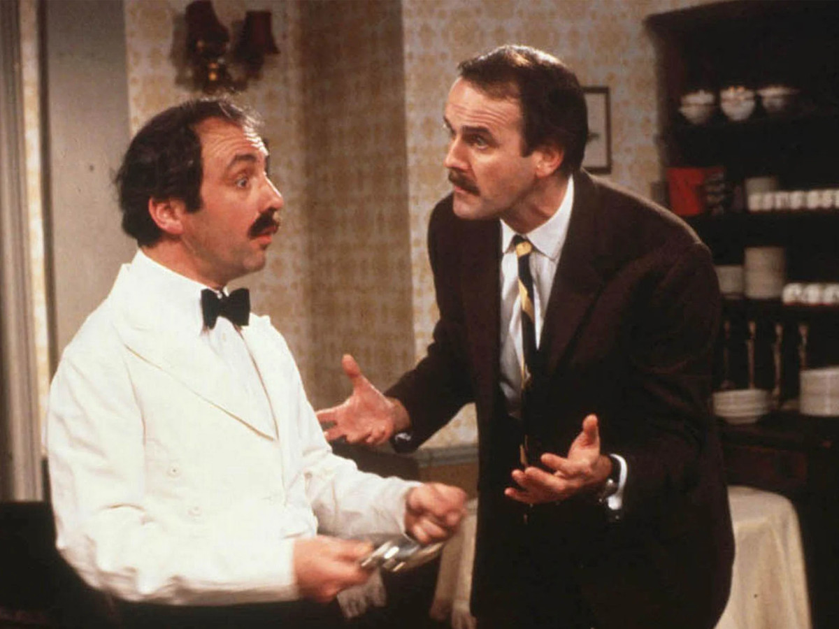 John Cleese, wearing a suit, yells animatedly at Andrew Sachs in a waiter’s outfit, in the dining room in Fawlty Towers.