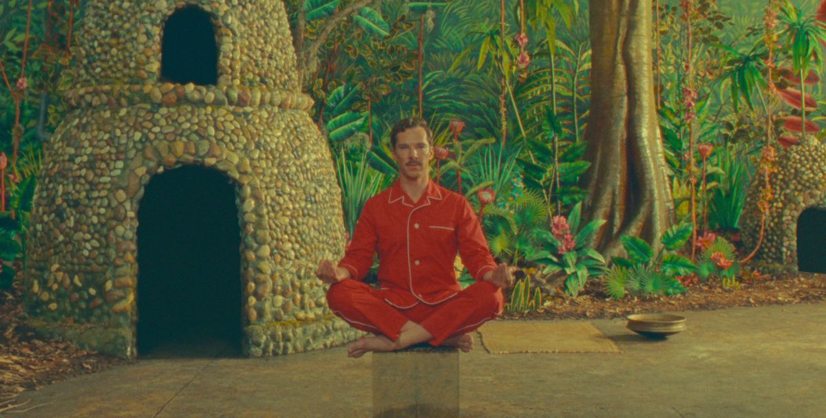 Henry Sugar (Benedict Cumberbatch, in red pajamas) sits crosslegged on a mostly transparent base designed to make him look like he’s levitating, in front of a stage-play-like backdrop of a green jungle and Indian-style forest hut in Netflix’s short The Wonderful Story of Henry Sugar