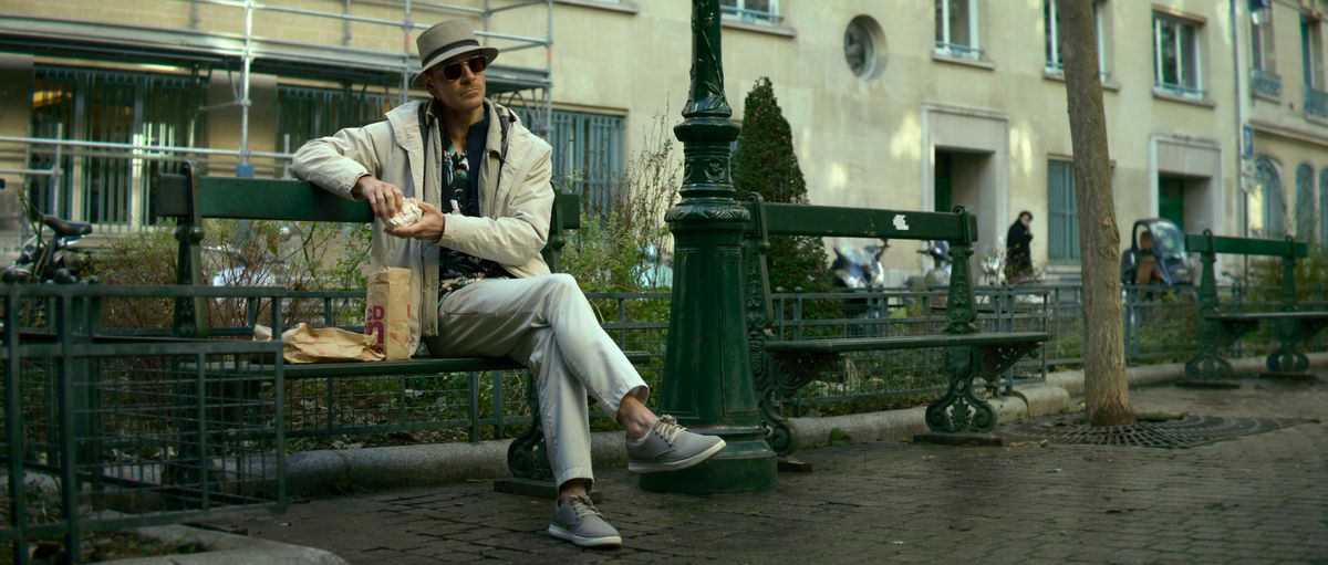 Michael Fassbender som Mördaren, sittande avslappnat på en bänk, klädd som en tysk turist