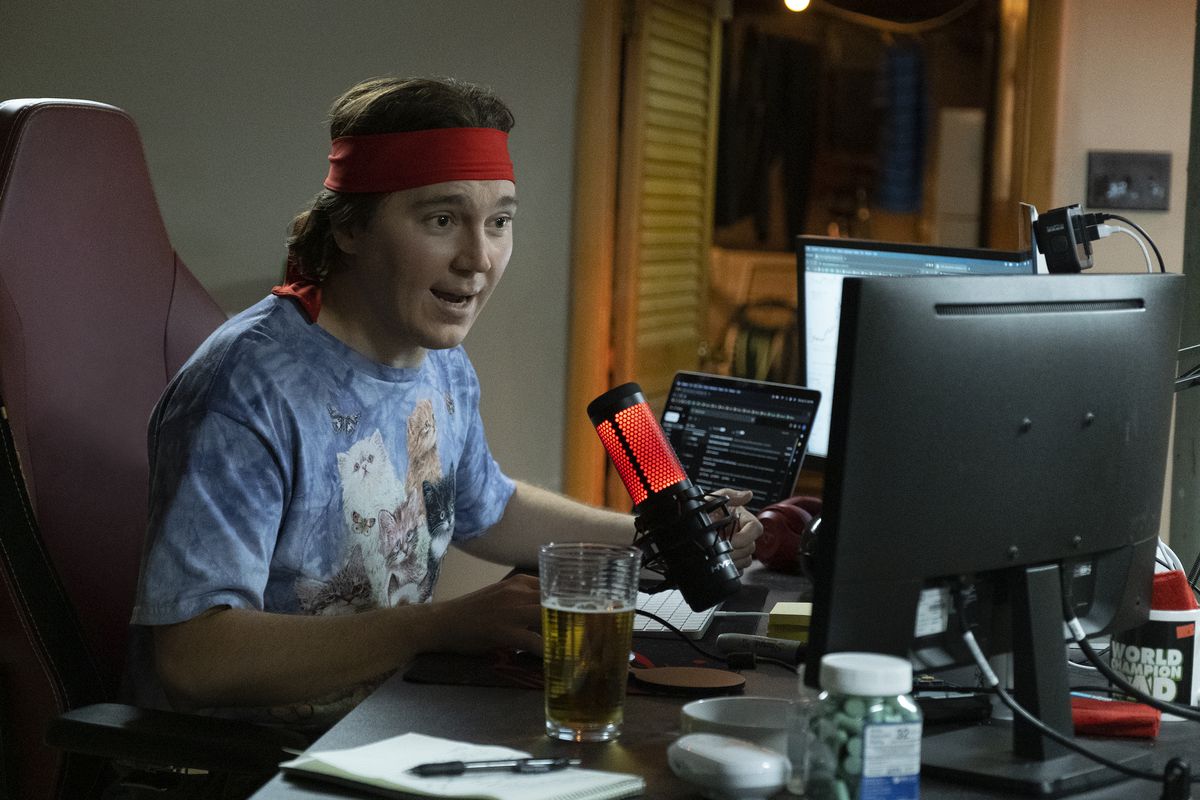 Paul Dano som Keith Gill, alias Roaring Kitty, sitter vid en bank av monitorer vid sitt skrivbord med en glödande mikrofon framför sig, klädd i en ny T-shirt med fluffiga kattungar och en röd bandana bunden runt hans huvud i Dumb Money