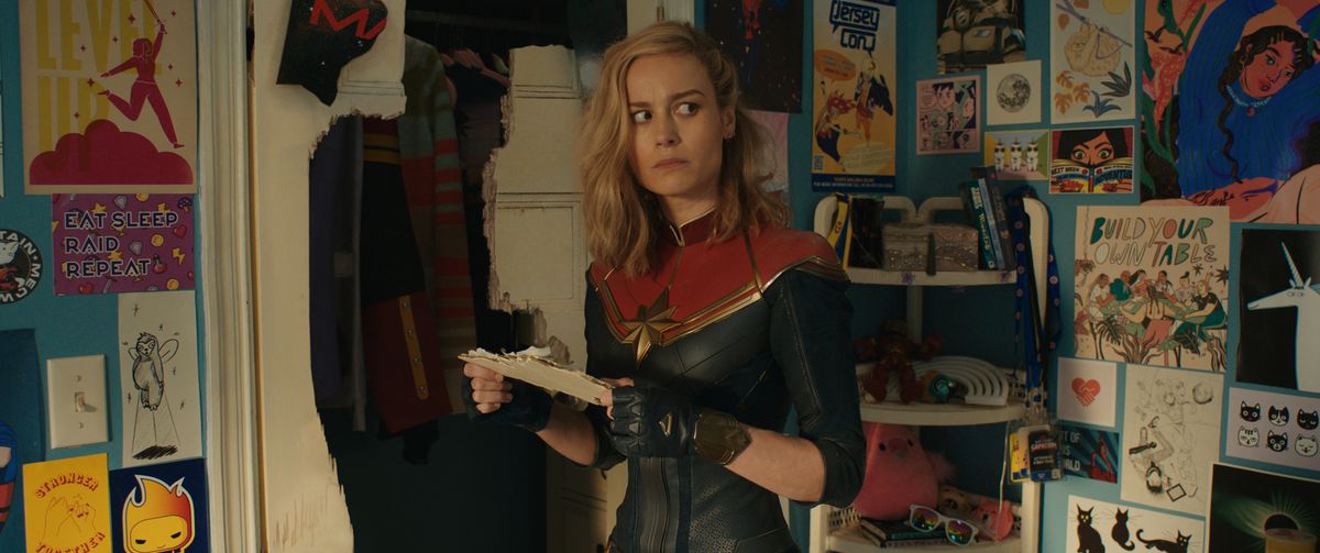 Kapten Marvel (Brie Larson) står i Kamala Khans sovrum bredvid en krossad garderobsdörr och ser sig oroligt omkring på alla tecknade bilder av sig själv, i Marvel Cinematic Universe-filmen The Marvels